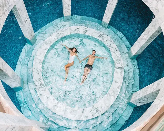 Une vue plus rapprochée d’en haut montre des personnes se reposant dans un bassin circulaire bleu pâle, encadré de motifs angulaires en marbre.