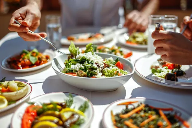 Une table garnie de salades colorées et de verres, avec des mains se tendant vers les plats.
