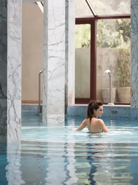 Une femme se repose dans une piscine intérieure turquoise, près de hautes fenêtres et de colonnes de marbre.