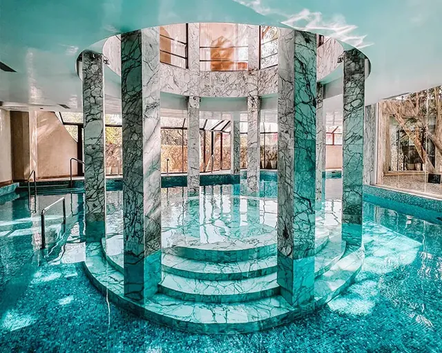 Une piscine intérieure turquoise entoure une structure centrale habillée de marbre, sous une haute verrière.