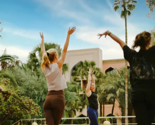 Des personnes lèvent les bras pendant une pratique de yoga en plein air sur une terrasse entourée de palmiers.