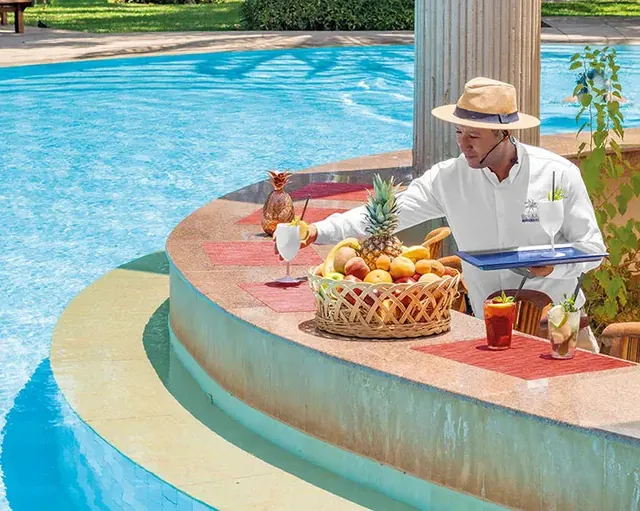Une personne en tenue blanche prépare des boissons fraîches à un comptoir arrondi, près d’une piscine extérieure.