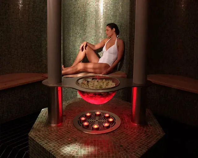 Une femme est assise sur un siège de pierre dans une salle de spa chaleureuse, face à un bassin éclairé en douceur.