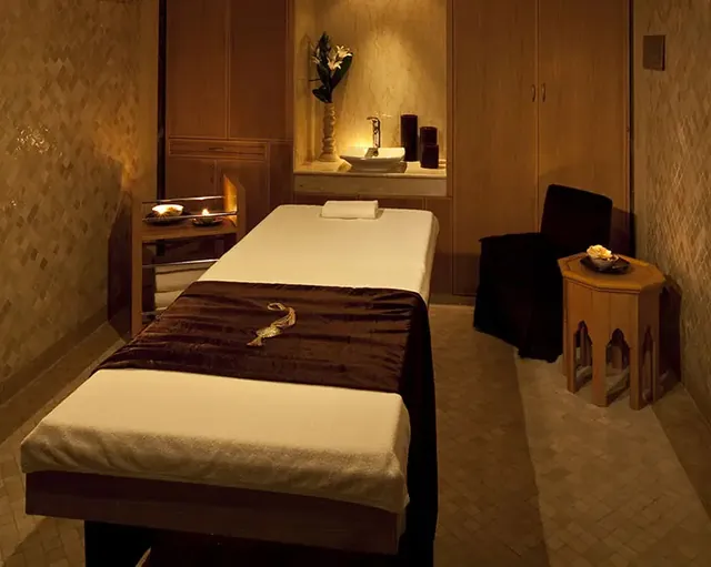 Une salle de soins à la lumière douce, avec des murs ocre, une table de massage et des meubles en bois foncé.