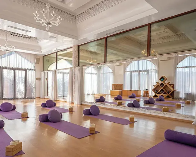 Des tapis, des briques et des bolsters violets sont disposés sur le parquet d’une salle lumineuse aux fenêtres cintrées.