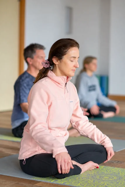 Une femme en veste rose est assise en tailleur, les yeux fermés et les mains sur les genoux, avec d’autres participants derrière elle.