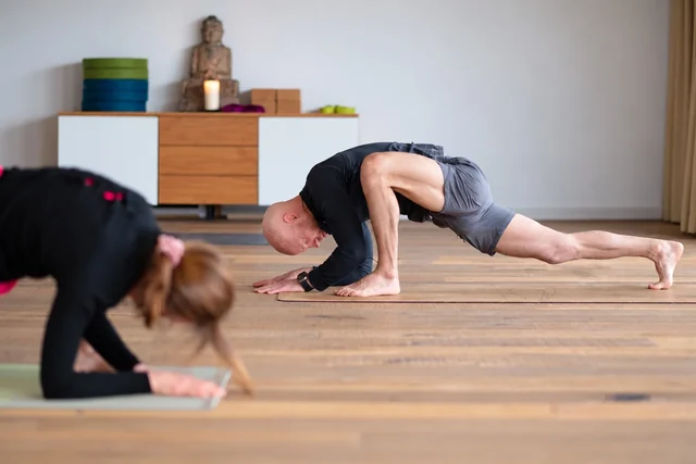 Un homme abaisse le buste dans une fente profonde, les avant-bras près du tapis, tandis que d’autres participants s’étirent à ses côtés.