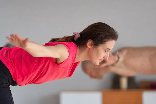 Une femme en rose se penche en avant, les bras tendus sur les côtés, tandis qu’un professeur de yoga fait une démonstration derrière elle.