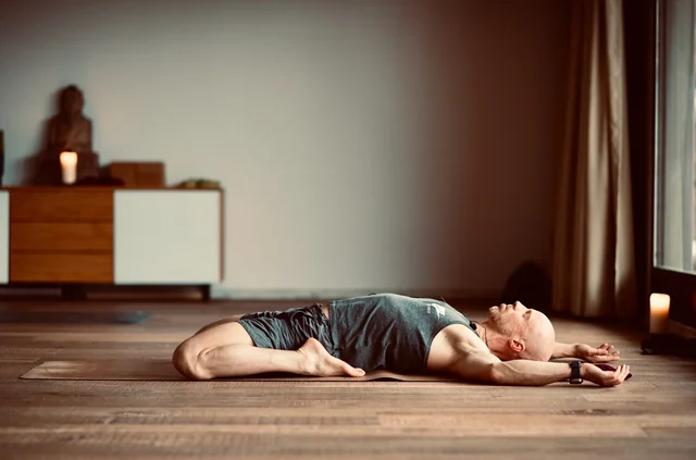 Un homme est allongé sur un tapis de yoga, les genoux repliés et les bras étirés au-dessus de la tête, près d’une fenêtre et de bougies allumées.