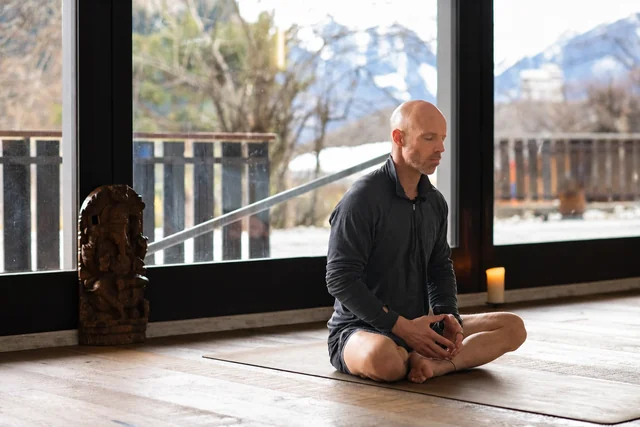 Un homme est assis en tailleur, les yeux fermés, sur un tapis de yoga près de grandes fenêtres donnant sur les montagnes enneigées et les arbres.