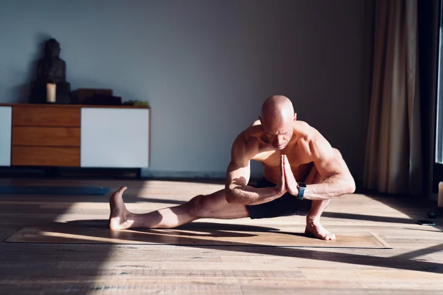 Un homme se tient en fente latérale profonde, une jambe tendue et les paumes jointes, dans la lumière du soleil sur le parquet du studio.
