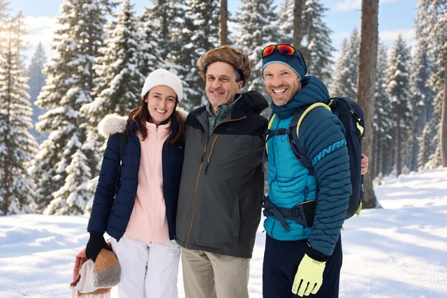 Trois randonneurs souriants en vêtements d’hiver se tiennent côte à côte parmi des conifères enneigés, dans une lumière douce.