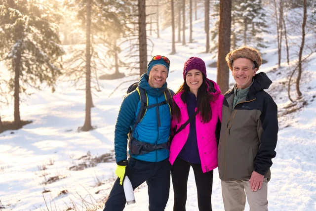 Trois personnes sourient en vêtements d’hiver sur un sentier forestier enneigé, dans la lumière chaude filtrant entre les arbres.