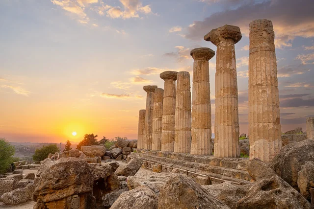A row of weathered stone temple columns rises above scattered ruins as the setting sun lights the clouds in gold.