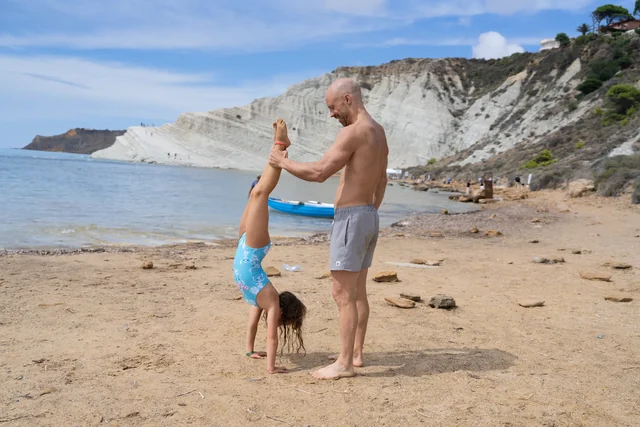 A man supports a child by the ankles as she holds a handstand on a sandy beach, with pale cliffs beside the sea.