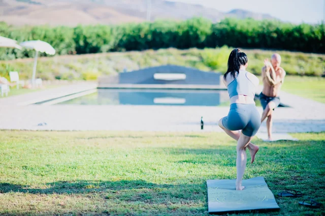 Two people balance in eagle pose on yoga mats on a garden lawn, with a swimming pool and hills behind them.