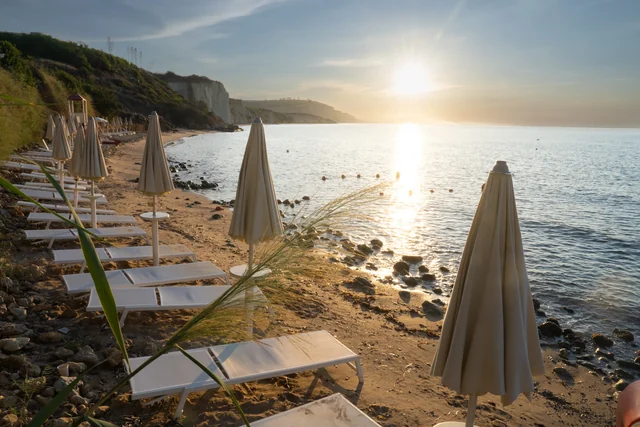 Golden sunlight reflects across a calm sea beside a sandy beach lined with loungers, closed umbrellas and pale cliffs.