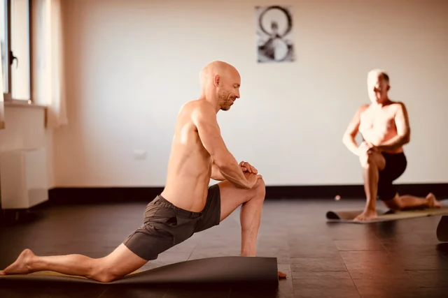 Two men practise kneeling low lunges on yoga mats in a softly lit room, with their hands resting on their front knees.