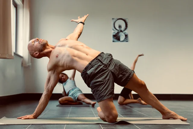 A man demonstrates a kneeling side bend with one hand on the floor and the other raised during a yoga class.