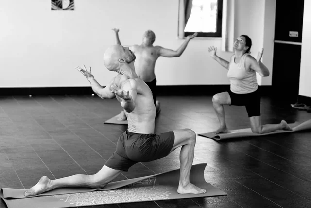 A black-and-white view of a teacher and participants practising low lunges with arms open in a yoga studio.