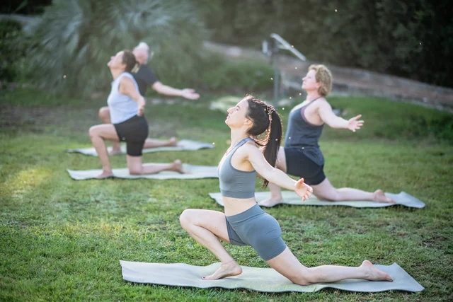 A woman opens her arms in a low lunge on a garden lawn while other participants stretch on yoga mats behind her.