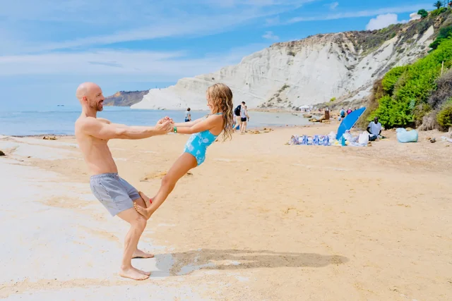 A man and child hold hands and lean away from each other in a playful partner balance on a sunny Italian beach.