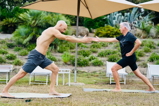 Two men face each other in standing lunges, reaching their front hands together on yoga mats in a green garden.