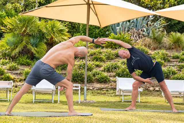 Two men lean into mirrored standing side bends and touch their raised hands above them during a garden yoga practice.