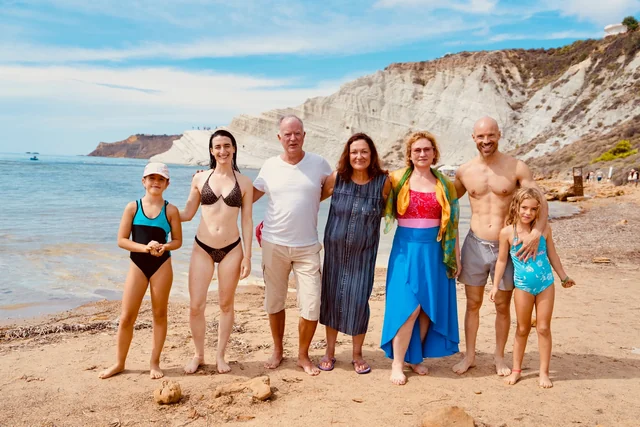 Seven people smile together for a group portrait on an Italian beach, with calm blue water and white cliffs behind them.