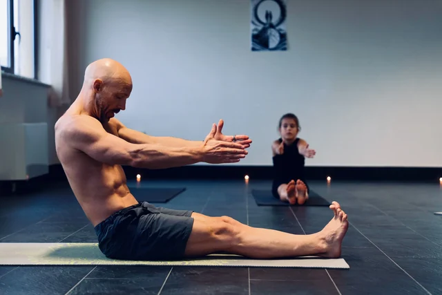A man sits upright with straight legs and reaches both arms forward during a yoga class, with a child following behind him.