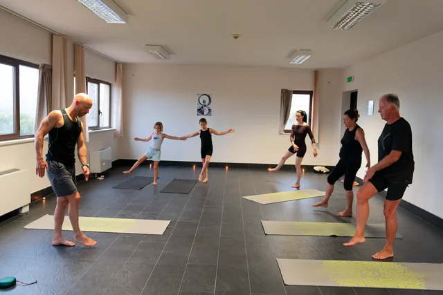Adults and children practise standing balances with one leg lifted during a group yoga class in a bright tiled room.