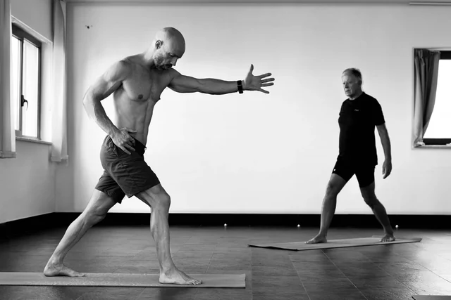 Black-and-white view of two men practising standing lunges, with the man in front extending one arm forward.