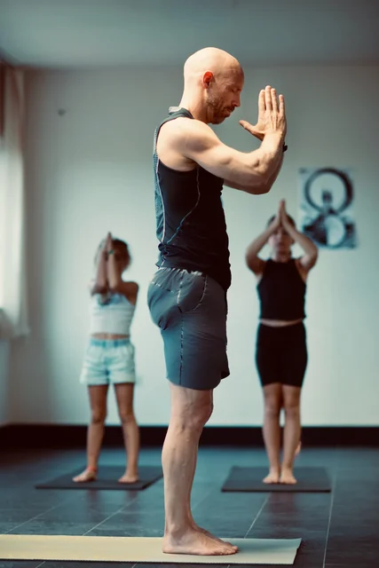 A man stands with palms together near his face while participants practise behind him in a yoga studio.