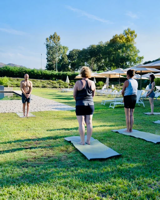 A teacher faces three participants standing with hands together on yoga mats in a sunny garden beside a pool.