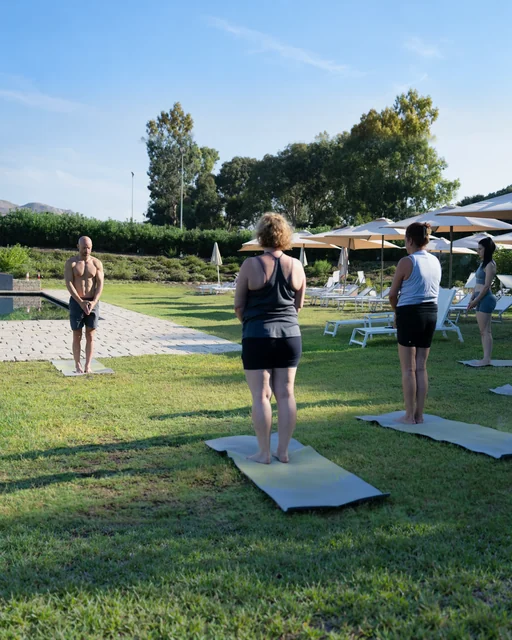 A teacher faces three participants standing with hands together on yoga mats in a sunny garden beside a pool.