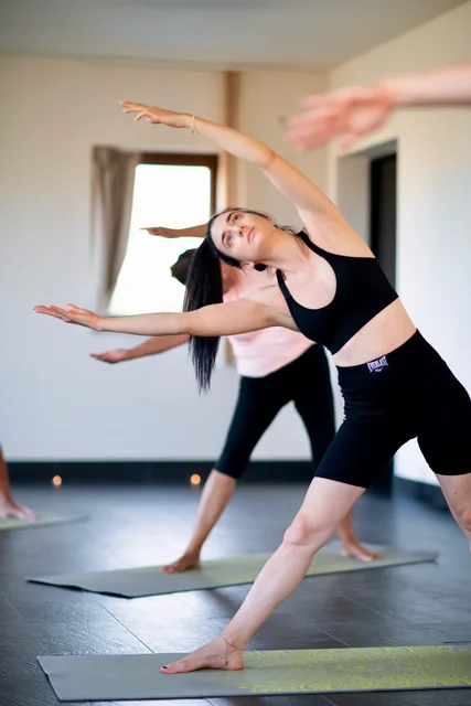 A woman leans sideways with both arms extended during a standing yoga class, with other participants practising behind her.