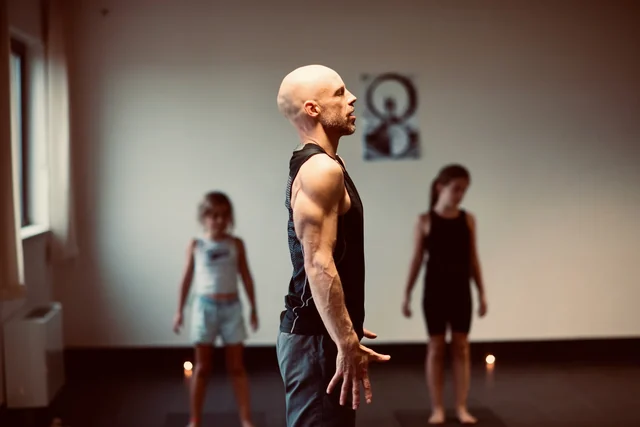 A man stands at the front of a yoga room with his arms relaxed, while two children practise behind him near the wall.