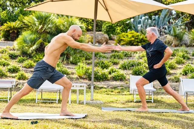 Two men reach toward each other in standing yoga lunges under a large garden umbrella, with bright sun on the lawn.