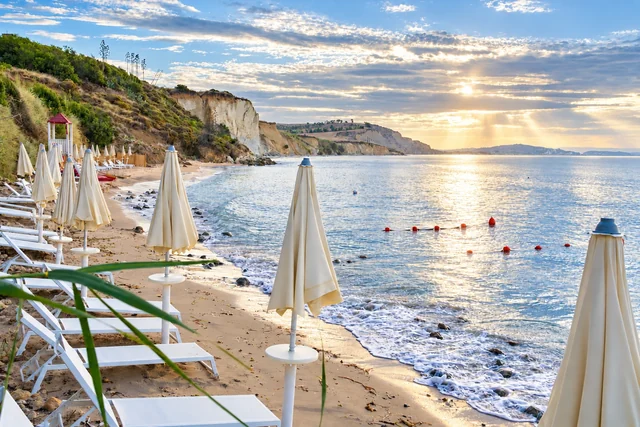 Sunrise shines through scattered clouds over an Italian beach, with white cliffs, closed umbrellas and sunlit water.