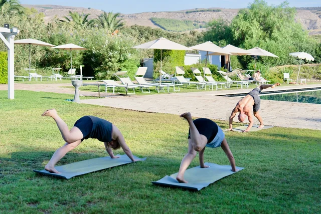 Three people practise three-legged downward dog on a shaded lawn beside a pool, sun loungers and parasols.
