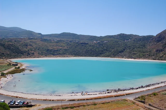 A turquoise lake curves around a pale shore below green hills, with a few people walking beside the water.