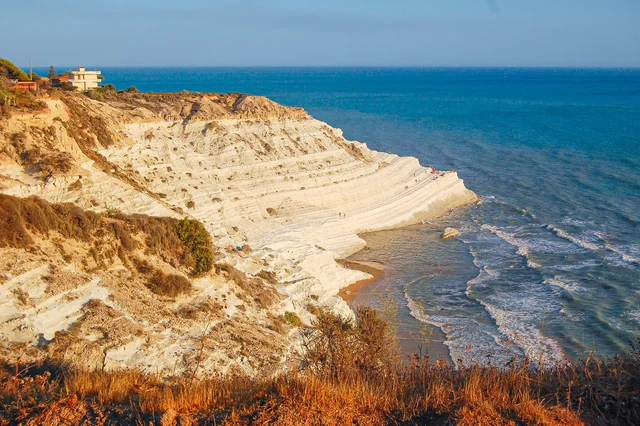 Pale, layered coastal cliffs curve down toward the blue sea, with warm sunlight illuminating the rocky shore.