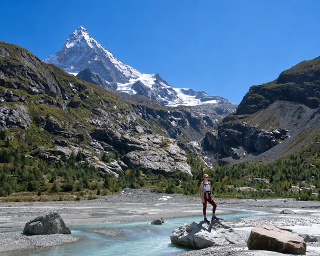 A hiker pauses on a river boulder beneath a tall snowy mountain, framed by rocky slopes, glacier ice and a clear blue sky.