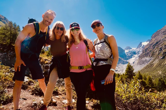 Four smiling hikers pose together on a sunny mountain trail, with steep rocky slopes and a glacier behind them.