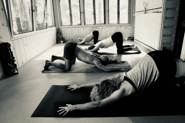Black-and-white view of a yoga group kneeling in Puppy pose with their hips lifted and arms stretched forward along their mats.