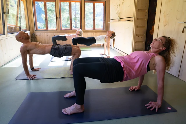 A group lifts their hips into reverse tabletop with bent knees and hands behind them, practising in a sunlit wooden yoga room.