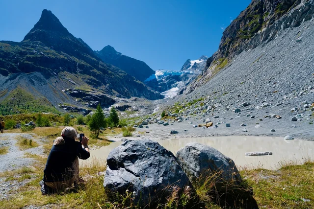 A seated hiker photographs a glacier from beside a large boulder, with pale water and rugged alpine peaks ahead.