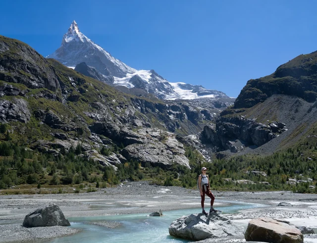 A hiker stands on a rock beside a braided alpine river beneath a pointed snowy summit, with a glacier across the valley.