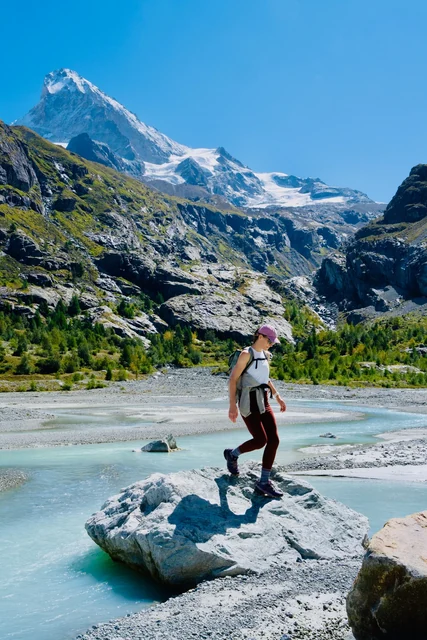 A woman with a backpack stands on a rock in a turquoise alpine stream, framed by rugged peaks and a glacier.