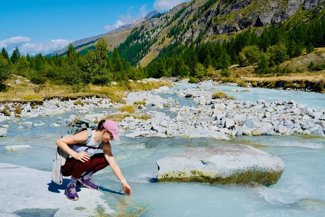 A woman crouches on a rock and reaches toward a turquoise alpine stream, with boulders and green slopes behind her.